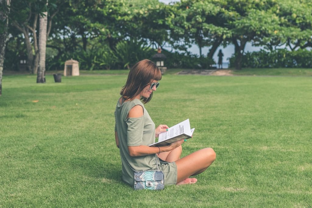 A woman with sunglasses reading a book in a park considering going back to school to become a wellness coach