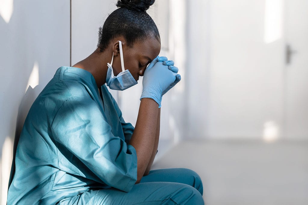 Nurse Burnout: Causes, Statistics, and Warning Signs 9 A burned out nurse squats in a hallway outside of a hospital room and holds her hands to her head.