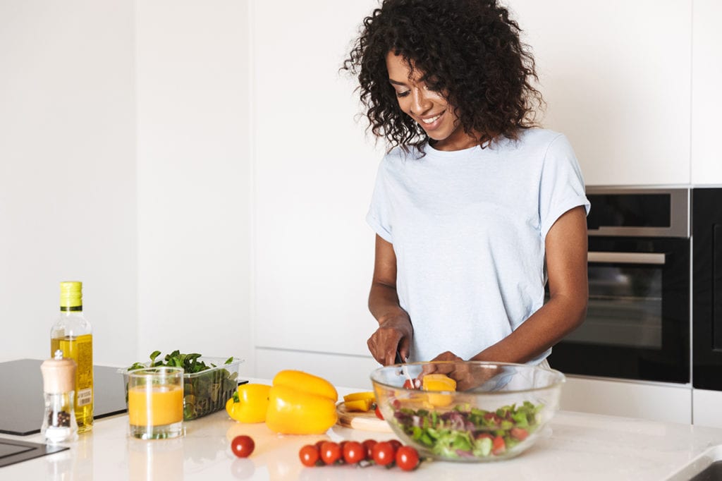 A happy woman chops vegetables for a salad at home after prioritizing her well-being to manage work stress and prevent job burnout.