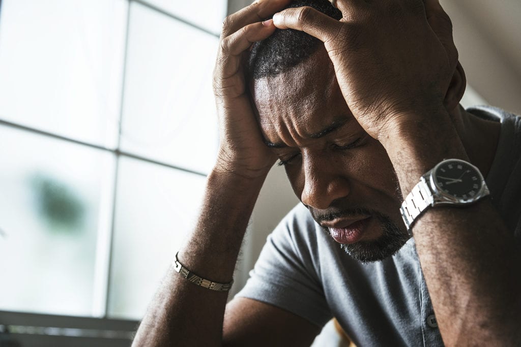 A stressed out man holds his head in his hands in front of a window. Ongoing stress at work can become dangerous job burnout over time.