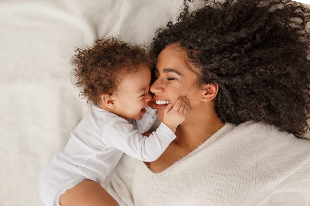 A mom and her baby in bed laughing and snuggling.